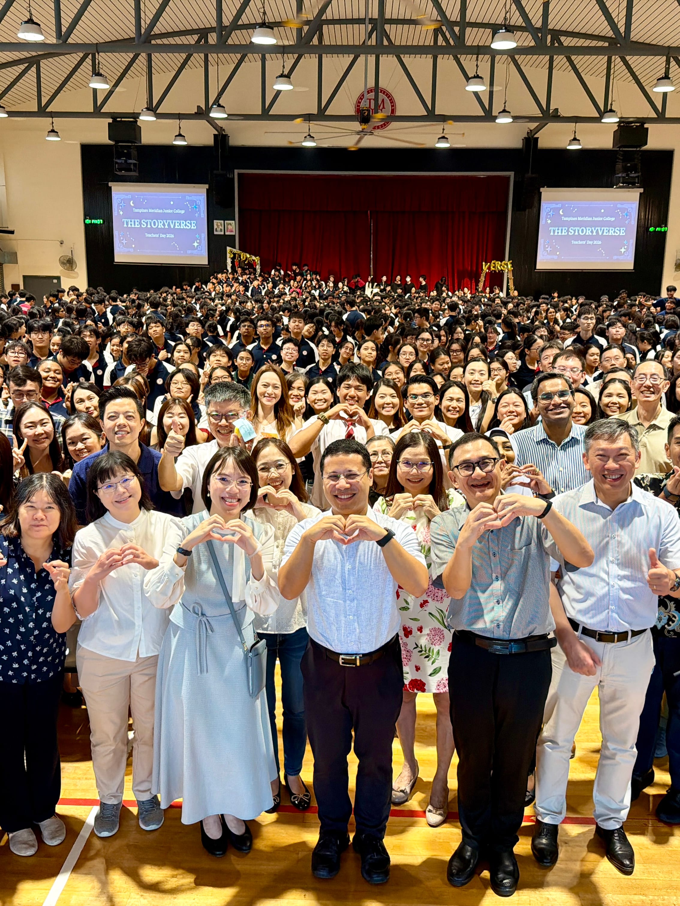 A large group photo of the entire college community — students, teachers, and staff — filling a school hall, with those in the front row making heart gestures with their hands. Two screens in the background display "The Storyverse – Teachers' Day 2026."
